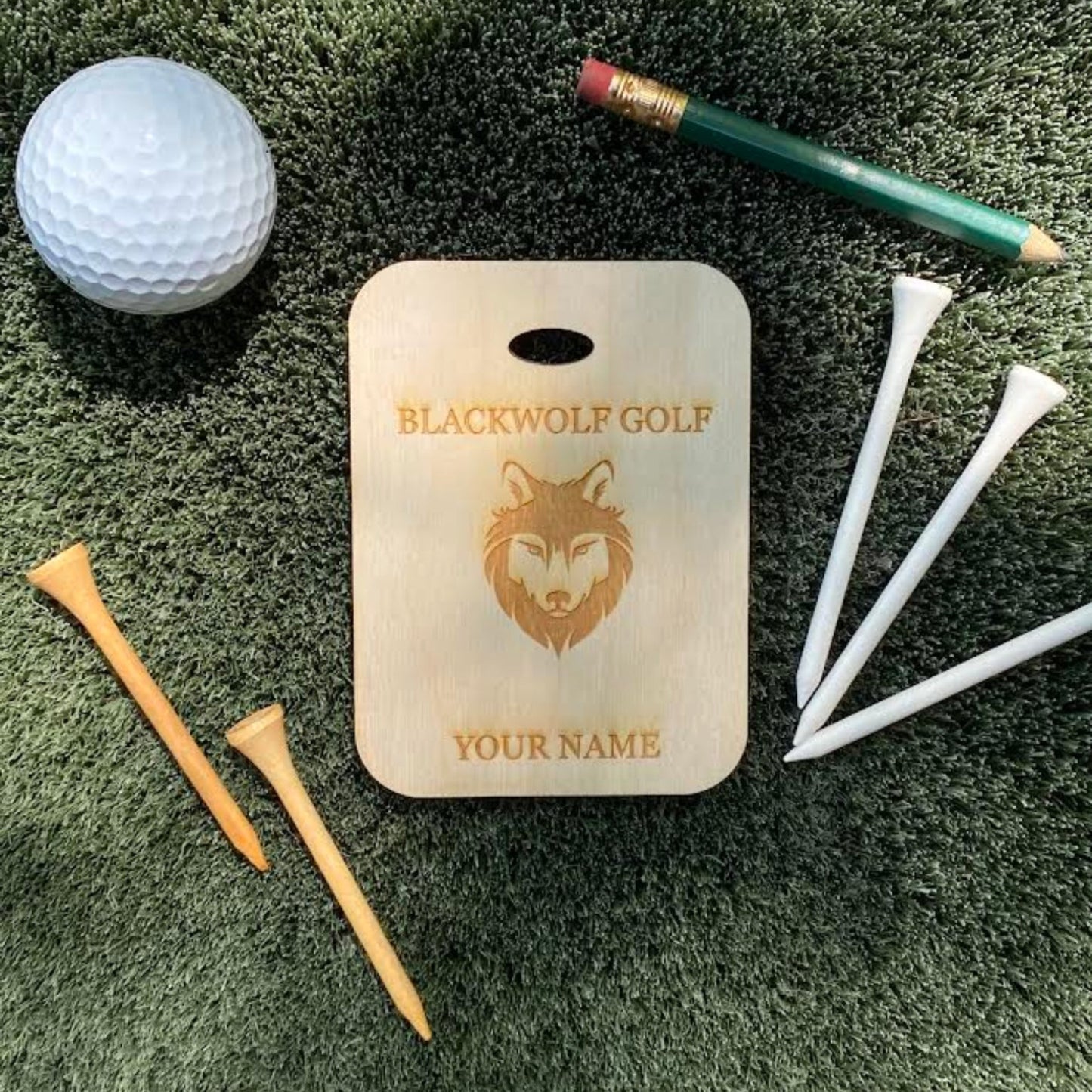 Golf ball, golf tees, pencil, and a 'Blackwolf Golf' branded bag tag on a grassy surface.